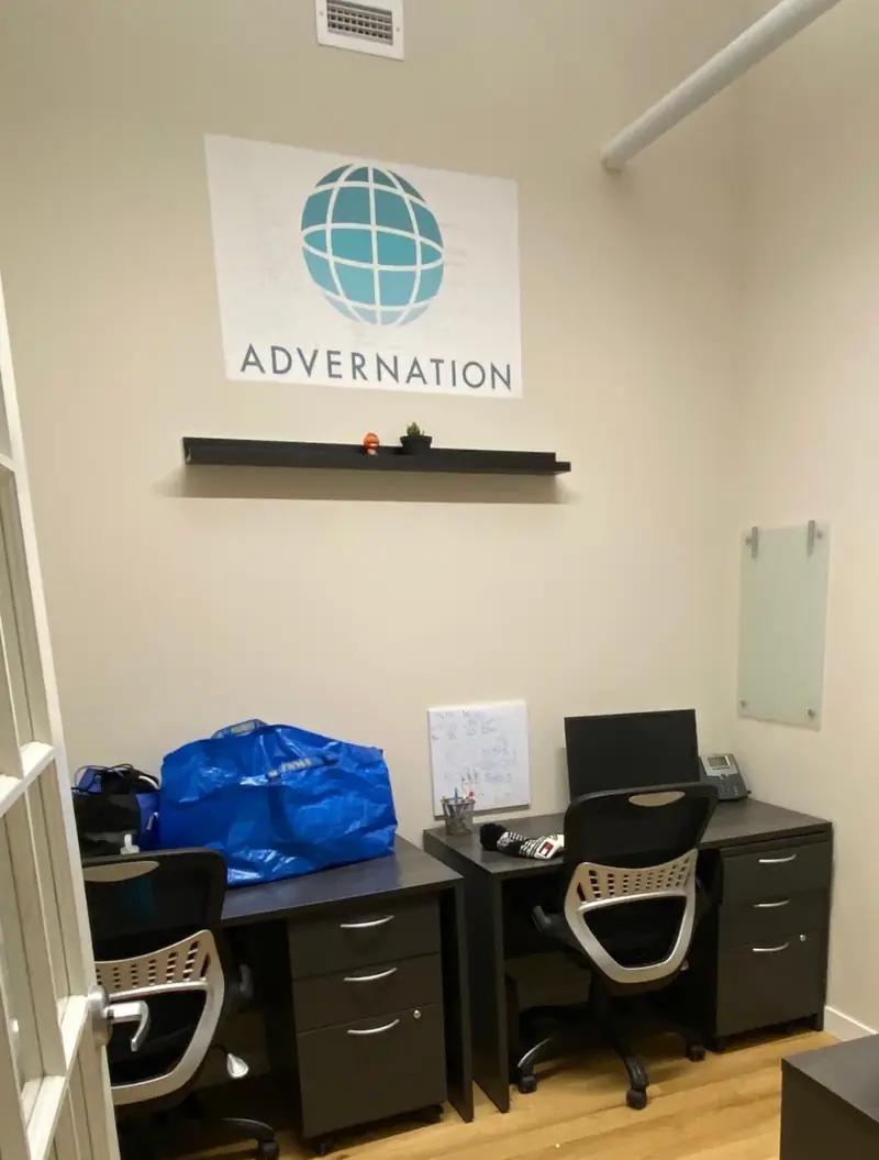 Two dark desks with office chairs under a wall sign showing a teal globe and the name Advernation, on a wooden floor