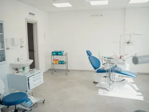 A bright dental treatment room with a blue chair, a stool, a sink and trolleys