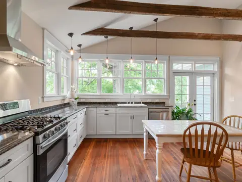 A bright kitchen with white cabinets, a wood floor, ceiling beams and a small table with chairs