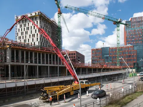 A building site beside a road with two tower cranes, a red concrete pump and a half-built block