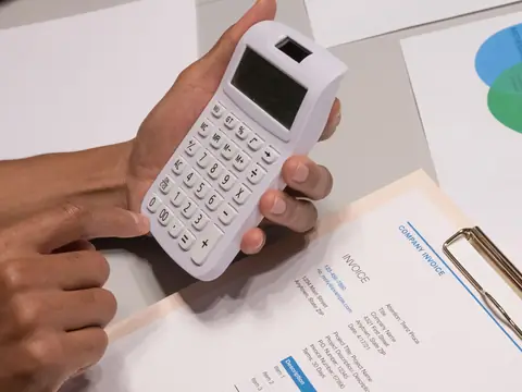 A hand holding a white calculator over a printed invoice on a clipboard