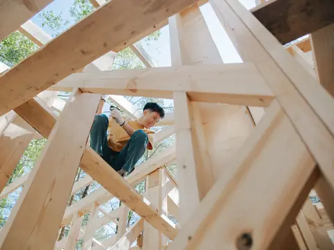 A carpenter sitting high in the timber frame of a house under construction, seen from below