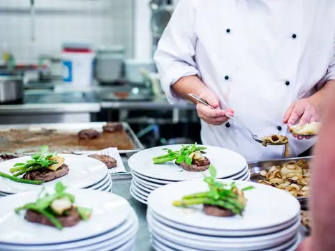 A chef's hands finishing rows of plated dishes on a steel counter