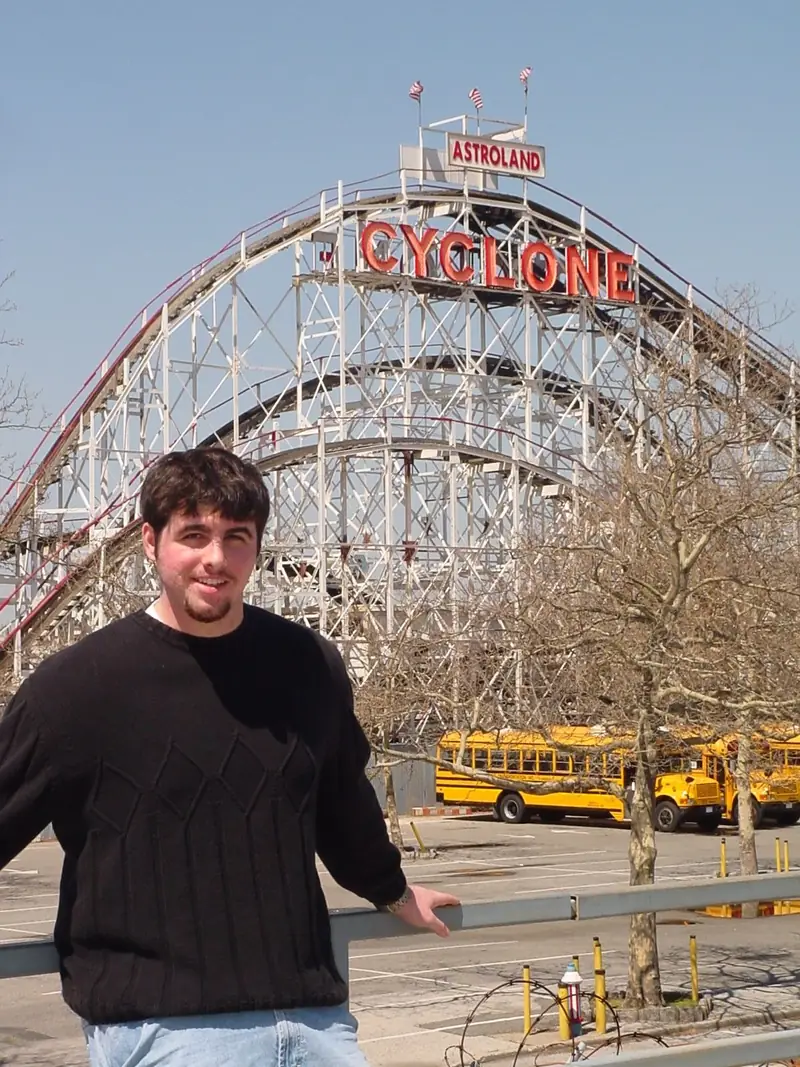 A young Christopher Maddaloni in a black sweater leaning on a rail in front of the Cyclone roller coaster at Coney Island