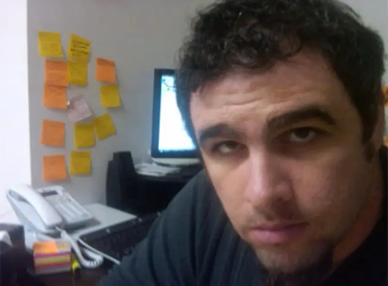 Christopher Maddaloni close to the camera at an office desk, with a desk phone, a monitor and sticky notes on the wall behind him