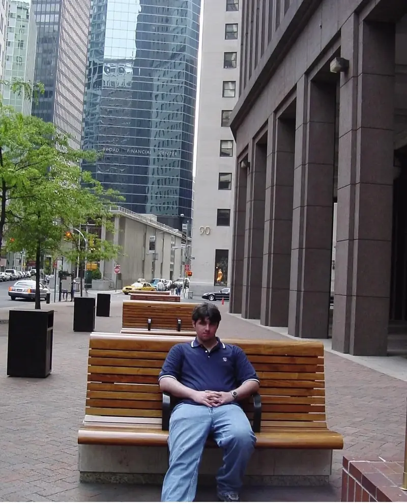 A young Christopher Maddaloni sitting on a wooden bench on a paved plaza between office towers, with the number 90 on a building behind him