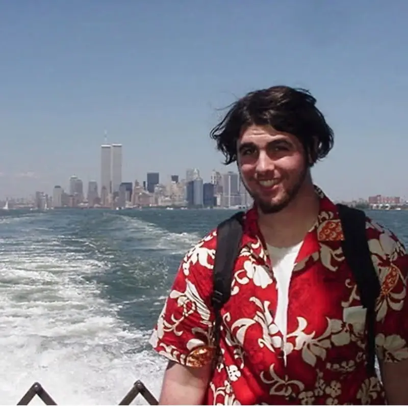 A young Christopher Maddaloni in a red flowered shirt at the stern of a ferry, with the boat's wake and the Lower Manhattan skyline behind him