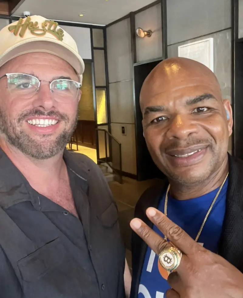 Christopher Maddaloni in a cream cap and glasses in a selfie with Davinci Jeremie, who holds up two fingers and wears a large ring