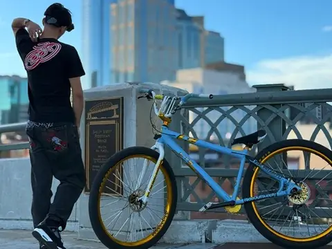 Christopher Maddaloni Jr. seen from behind beside a light blue bike with gold wheels on a bridge, a glass tower behind him