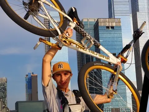 Christopher Maddaloni Jr. in a tan cap, holding a white bike with gold wheels over his head in front of glass towers