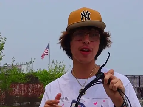 Christopher Maddaloni Jr. in a tan cap and glasses, talking to the camera and holding up a set of bike brake cables