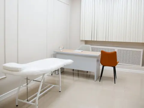 A white consulting room with an examination couch, a desk and an orange chair