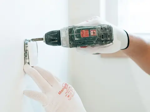 A gloved hand holding a cordless drill to a socket box on a white wall
