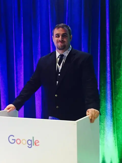Christopher Maddaloni in a dark blazer standing behind a white lectern with the Google logo on it, in front of blue and green stage curtains