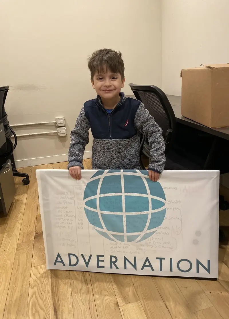 A small boy in a grey and navy pullover standing behind a white banner with a teal globe and the name Advernation, in an office