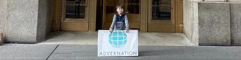A small boy standing on a sidewalk behind a white banner with a teal globe and the name Advernation, in front of brass doors