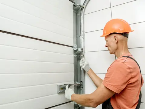 A worker in an orange hard hat tightening the track of a white sectional garage door