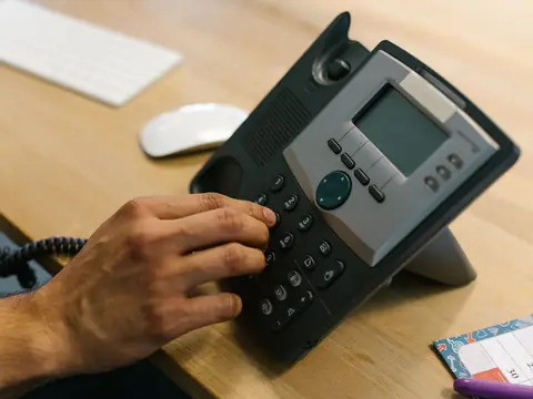 A hand pressing the keys of a black desk phone on a wooden desk