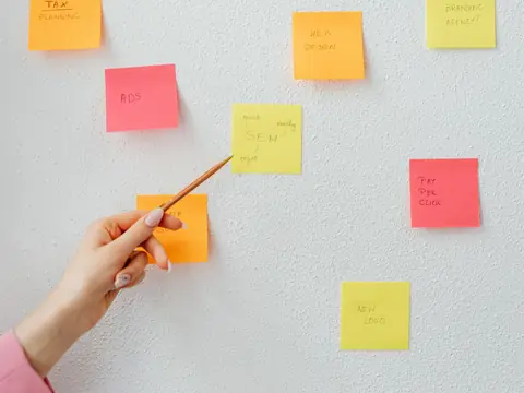 A hand pointing a pencil at coloured sticky notes on a white wall