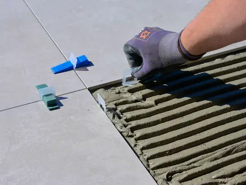 A gloved hand setting a large floor tile into combed adhesive, with blue levelling clips