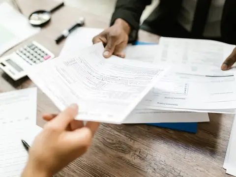 Hands passing printed documents across a desk with a calculator and a magnifying glass