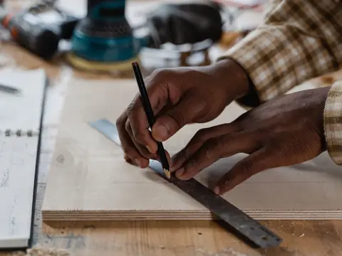 Two hands marking a board with a pencil against a steel square on a workbench