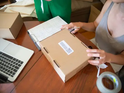 Hands taping a labelled cardboard parcel on a table beside a laptop