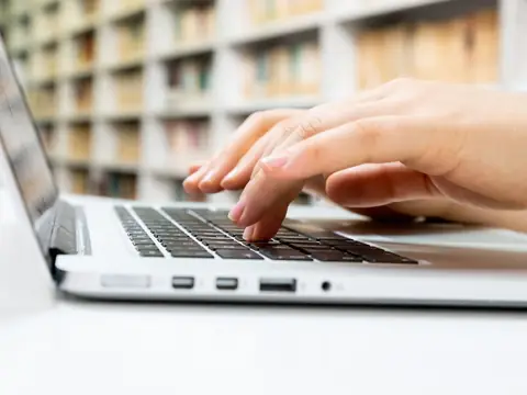 Two hands typing on a silver laptop, with bookshelves out of focus behind