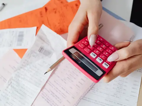 Hands working a pink calculator over a spread of receipts and invoices