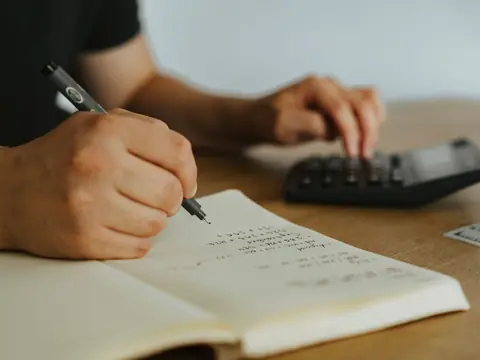 A hand writing sums in a notebook while the other hand presses the keys of a calculator
