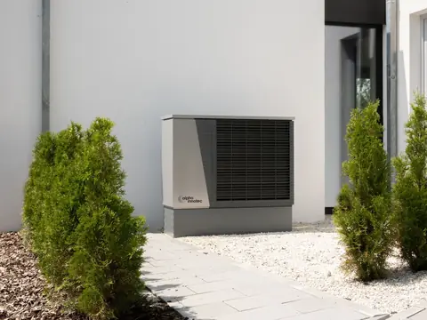 A grey heat pump unit standing on gravel beside a white house, between small conifers