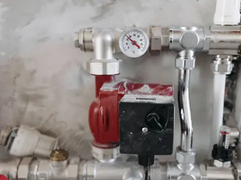 Chrome heating valves with a round pressure gauge and a red pump on a grey wall