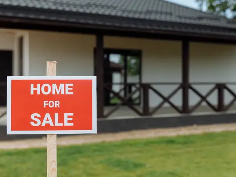 A red sign reading Home for sale on a post in the lawn in front of a house with a veranda