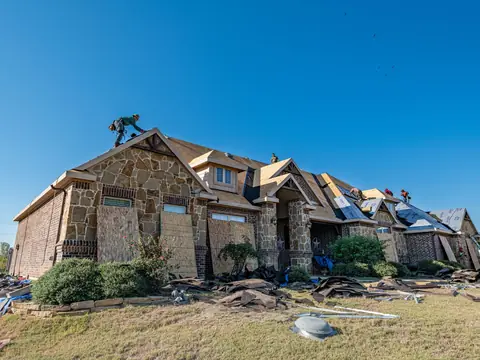 A stone house with its roof stripped for new shingles and a worker on the ridge, under a blue sky