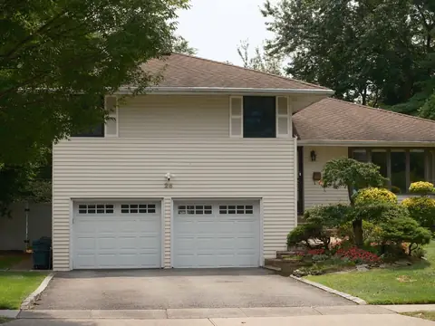 A two-storey house with cream siding, two white garage doors and a paved driveway, with trees around it