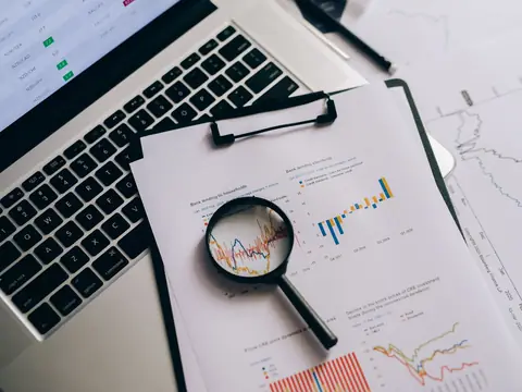 A magnifying glass lying on printed charts on a clipboard, beside an open laptop