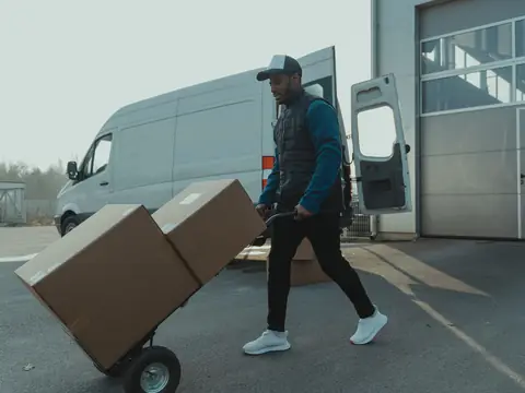 A man in a cap pushing a hand truck of cardboard boxes past a white van with its doors open
