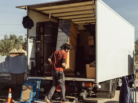 A man carrying a piece of furniture into the open back of a box truck already loaded with furniture and boxes