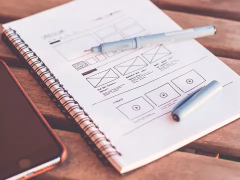 A spiral notebook with hand-drawn website layout sketches, two pens lying on it and a phone beside it