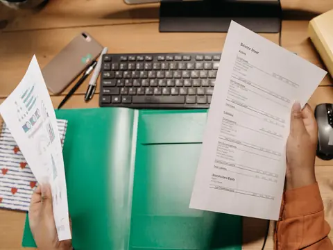Two hands holding printed pages side by side over a desk with a keyboard and a green folder