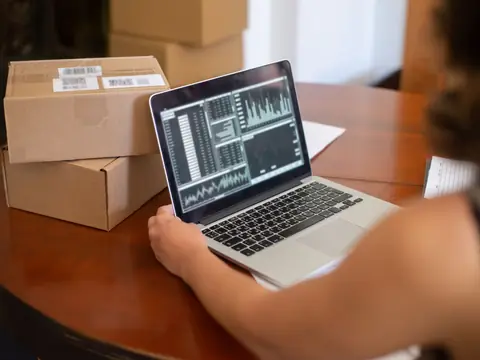 A person at a round table with a laptop showing charts, beside two labelled parcels, seen from behind