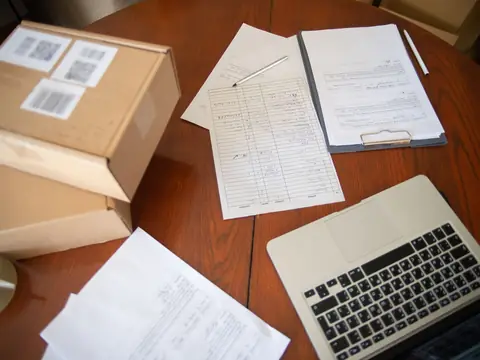 Cardboard parcels with labels, printed order sheets and a laptop on a wooden table, seen from above
