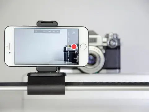 A white phone on a desk mount with its camera recording, and a film camera behind it