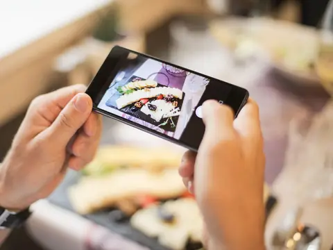Two hands holding a smartphone sideways to photograph a plate of food on a table
