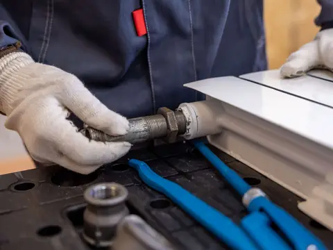 Gloved hands tightening a pipe fitting on a white radiator, with blue pipe wrenches lying below