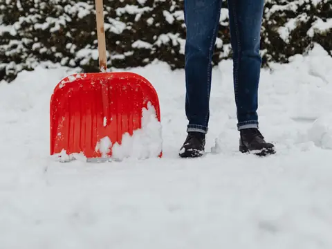 A red snow shovel standing in deep snow beside a person's legs