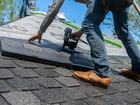 A roofer bending to fix dark shingles to a roof with a nail gun, under a blue sky