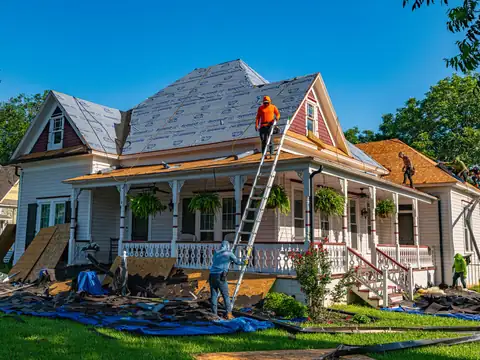 A wooden house with a porch being re-roofed, a roofer on the roof and another on a ladder