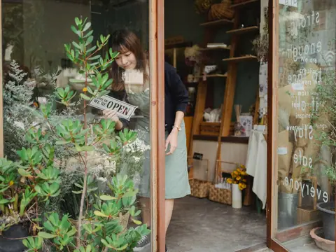 The open door of a small shop with plants outside and a sign hanging on the glass