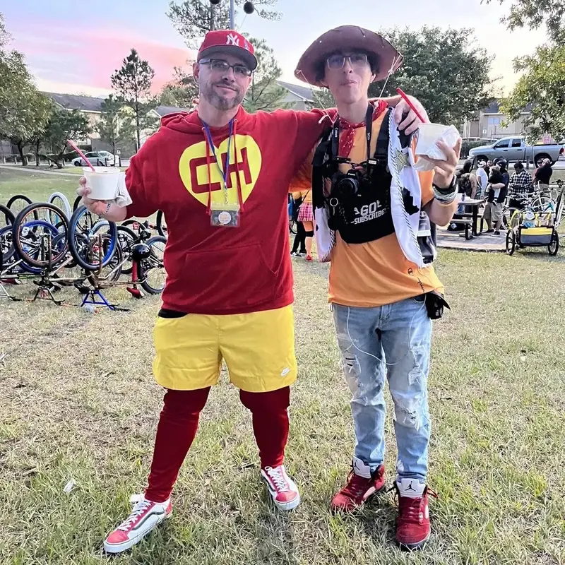 Two people in costume on the grass at a bike event, one in a red hooded suit with yellow shorts, with rows of bicycles behind them